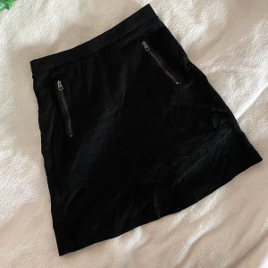 Svart kortkjol från Marc O'Polo - Säljer en svart kortkjol från Marc O'Polo med två snygga dragkedjor framtill. Kjolen har en enkel och stilren design som passar perfekt till många olika outfits. Midja: 33cm, Längd: 47cm