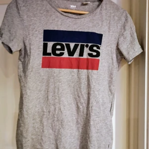 Grå Levi's t-shirt med logga - Snygg grå t-shirt från Levi's med klassisk stor logga i blått, svart och rött framtill. T-shirten har rund halsringning och är kortärmad. Tillverkad i mjuk bomull för en bekväm känsla. Sparsamt använd 