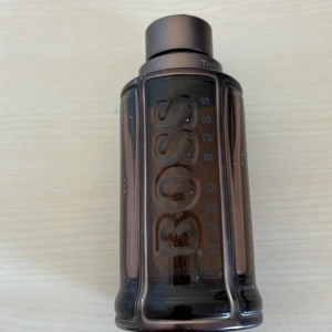 BOSS The Scent Absolute Eau de Parfum 100 ml - En elegant herrparfym från Hugo Boss, The Scent Absolute, i en mörkbrun glasflaska med metallisk finish. Flaskan rymmer 100 ml och har en robust, maskulin design med tydlig logotyp framtill.