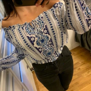 Blåvit offshoulder blus med mönster - Säljer en blåvit offshoulder blus med långa ärmar och elastisk midja. Blusen har ett vackert blått mönster med knytning framtill och volangkant upptill. Perfekt för en somrig och bohemisk look.