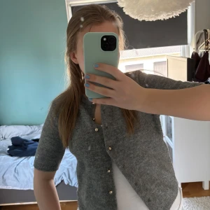 Grå kortärmad kofta från H&M - Mysig grå kofta från H&M med korta ärmar och rund halsringning. Tröjan har fina guldfärgade knappar framtill och är stickad i ett mjukt material. Perfekt att slänga över en topp för en enkel och stilren look.