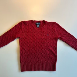 Röd kabelstickad tröja från Polo Ralph Lauren - Riktigt snygg röd kabelstickad tröja från Polo Ralph Lauren med rund halsringning och klassisk broderad logga på bröstet. Tröjan har lång ärm och ribbade muddar vid ärmslut och nederkant. Perfekt för lager-på-lager-stil. Passar för cirka 5-6 åringar ( XXS ) .