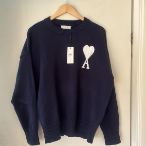 Mörkblå stickad tröja från Ami - Säljer en mörkblå stickad tröja från Ami med en vit broderad hjärtlogga och bokstaven A på bröstet. Tröjan har rund halsringning och långa ärmar. Perfekt för dig som gillar stilrena och trendiga plagg.