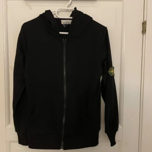 Svart hoodiejacka från Stone Island - Svart hoodiejacka från Stone Island med dragkedja framtill och klassisk logotyp-patch på vänster ärm. Jackan har huva och två fickor framtill. Perfekt för en stilren och avslappnad look. Storlek S