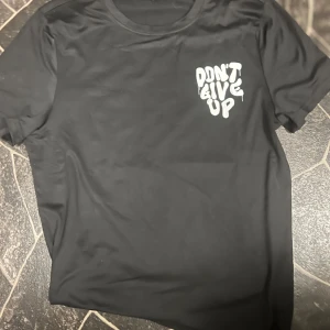 Svart t-shirt med tryck 'Don't Give Up' - Cool svart t-shirt med stort vitt och rött tryck både fram och bak där det står 'Don't Give Up', 'Never Stop Trying' och 'Stay Positive'. Rund halsringning och korta ärmar. Perfekt för dig som gillar statement-plagg.