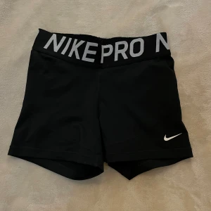 Svarta Nike Pro shorts - Svarta Nike Pro shorts med bred resår i midjan där det står 'NIKE PRO' i vitt. Tight passform och korta ben, perfekt för träning. Materialet är syntet och har en liten vit Nike-logga på ena benet.