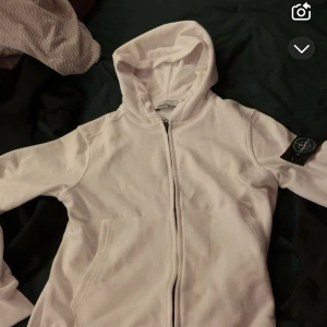 Vit hoodie från Stone Island - Säljer en vit hoodie från Stone Island med dragkedja framtill och klassisk patch på ärmen. Tröjan har huva och två fickor framtill, perfekt för en clean och stilren look.
