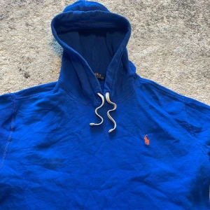 Blå hoodie från Polo Ralph Lauren - Säljer en klassisk blå hoodie från Polo Ralph Lauren med orange broderad logga på bröstet. Tröjan har huva med vita snören och en stor magficka. Perfekt för dig som gillar stilrena och sportiga plagg.