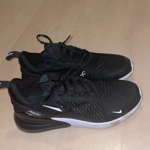  Air Max 270  - Säljer ett par svarta Nike Air Max 270 sneakers med vit sula och diskreta vita detaljer. Skorna har en sportig design med mesh-överdel och synlig Air Max-dämpning i hälen. Perfekta för dig som gillar stilrena och bekväma sneakers. Perfekt skick