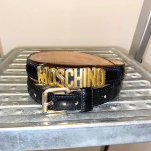 Skärp från Moschino - Ikoniskt smakt Moschino-bälte i svart läder med guldfärgade bokstäver. Kan även att användas som halsband.   Mått: 88 cm.   /   Spara skärmtid, besök våra butiker i Sthlm, Malmö & Gbg. New arrivals daily + studentrabatt i alla våra butiker! Hitta till din närmsta butik på humansecondhand.se.   HUMANA Second Hand erbjuder 14 dagars retur, även via plick. Returer skickas med PostNord. Vid retur kontakta support@plick.se.