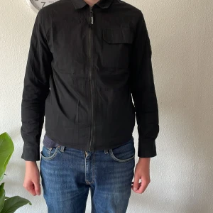 Svart overshirt från C.P. Company - Säljer en svart overshirt från C.P. Company med dragkedja framtill och klassisk krage. Jackan har en ficka på bröstet och en ikonisk linsdetalj på ärmen. Perfekt för lager-på-lager och stilren look.  några frågor