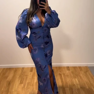 Blå långklänning från gina tricot - säljer en adoore liknande klänning. Säljer en blå långklänning med ballongärmar och djup v-ringning. Klänningen har knappar framtill, slits och ett blommigt mönster. Materialet har en lätt glansig finish och ger en elegant känsla. Helt ny med lappen kvar.💙