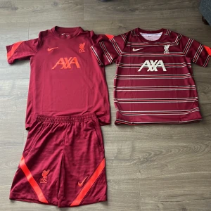 Liverpool Nike fotbollströjor  - Två Liverpool fotbollströjor från Nike i vinröd färg, båda med AXA och LFC-logga. Ena tröjan är helfärgad med röda detaljer på ärmarna, den andra har vita och svarta ränder. Båda är kortärmade och tillverkade i Dri-FIT material för bästa komfort.