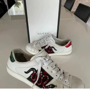 Gucci Ace sneakers med orm brodyr - Gucci Ace sneakers i vitt skinn med ikonisk röd och grön orm brodyr på sidan. Baksidan har metallicröd och metallicgrön detalj med ormskinnsmönster och Gucci-logga. Klassisk rund tå, snörning och platt sula.