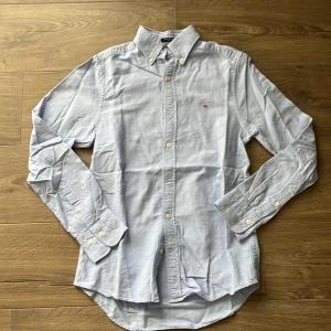 Ljusblå oxfordskjorta från GANT  - Snygg ljusblå oxfordskjorta från GANT i slim fit. Skjortan har klassisk button down-krage, broderad GANT-logga på bröstet och är tillverkad i mjuk bomull. Långärmad modell med knappar framtill och vid ärmslut. Perfekt för en clean och stilren look.