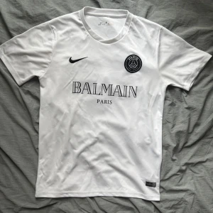 Vit PSG x Balmain fotbollströja Nike storlek S/M - Snygg vit fotbollströja från Nike med Paris Saint-Germain och Balmain Paris tryck på bröstet. Klassisk rund hals, korta ärmar och svart Nike-logga. Tillverkad i lätt och ventilerande Dri-FIT material som passar perfekt för träning eller match. Storlek Small men passar ven som en medium