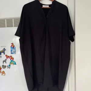 Svart kortärmad skjortklänning  från Femme Collection - Enkel och stilren svart skjortklänning från Femme Collection. Klänningen har lös passform, korta ärmar och v-ringad halsringning. Perfekt för dig som gillar minimalistisk stil och vill ha något bekvämt men snyggt.