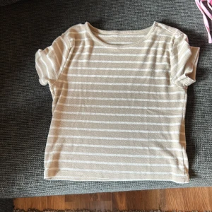 Beige och vit randig topp från 157 - Kortärmad topp från 157 i beige och vitt med horisontella ränder. Modellen är enkel med rund halsringning och mjukt, stretchigt material. Perfekt för en chill och avslappnad stil. Materialet är polyesterblandning för extra komfort.