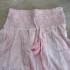 Ljusrosa långkjol med tofsar - Supersöt ljusrosa långkjol med smock i midjan och knytband med pärlprydda tofsar. Kjolen har ett diskret paisleymönster i vitt och är gjord i ett tunt, flowy material som känns perfekt för varma dagar.