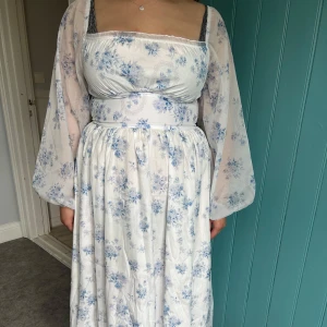 Vit och blå blommig långklänning Shein - Romantisk långklänning från Shein i vitt med blått blommönster. Klänningen har transparenta, vida långärmar och markerad midja. Materialet är glansigt och lätt, med dragkedja i ryggen och fyrkantig urringning. Perfekt för dig som gillar cottagecore-vibes.