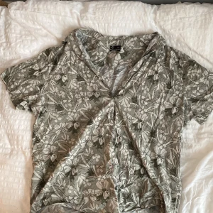 Blommig grön kortärmad skjorta C 2XL - Kortärmad skjorta från C i storlek 2XL med grön bas och stora vita blommor över hela tyget. Regular fit och mjukt bomullsmaterial, perfekt för dig som gillar avslappnad stil med lite extra mönster. Skjortan har v-ringning och känns riktigt chill. Sitter baggy 🫶🫶