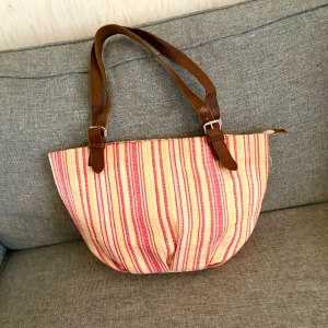 Randig axelväska från Don Donna - Säljer en randig axelväska från Don Donna med breda bruna skinnremmar och dragkedja upptill. Väskan har röd, beige och gul randig bomullsyta och rosa foder inuti. Perfekt för dig som gillar färg och vill ha en unik accessoar till vardagslooken.