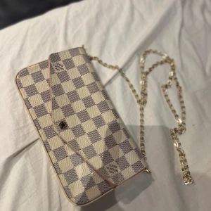 Louis Vuitton rutig axelväska med kedja - Väskan är äkta och du får den med box + en korthållare till. Säljer en snygg axelväska från Louis Vuitton i klassiskt rutigt mönster. Väskan är beige och grå med guldfärgad kedja som axelrem. Perfekt storlek för det viktigaste och stängs med tryckknapp. Kommer med originalbox och certifikat. Använt väskan två gånger! 