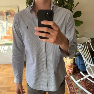 Randig skjorta från Ralph Lauren - Klassisk skjorta från Ralph Lauren med tunna blå/lila och vita ränder, broderad logga på bröstet och knappar framtill. Skjortan har lång ärm, button-down krage och en normal passform. Pris kan diskuteras, men den är i riktigt bra skick! 