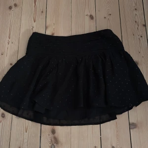 Svart kortkjol från Gina Tricot - Supersöt svart kortkjol från Gina Tricot med diskreta små prickar i glittrigt material. Kjolen har ett brett midjeband och är lätt utställd med volangig känsla. Perfekt för dig som vill ha en enkel men ändå lite extra kjol.