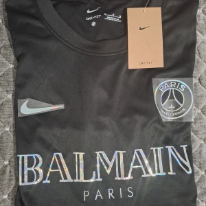 Svart Nike x Balmain Paris t-shirt - Svart t-shirt med holografiskt Balmain Paris-tryck framtill och Nike-logga. Crewneck och klassisk passform. Materialet är Dri-Fit, perfekt för en sportig look. Paris Saint-Germain-märke medföljer. Snygg och trendig för dig som gillar streetwear.