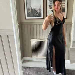 Svart långklänning med slits - Snygg svart långklänning i satinliknande material med smala axelband som korsas i ryggen. Klänningen har en djup v-ringning, cut-out detalj under bysten och hög slits på ena sidan. Perfekt för dig som vill sticka ut med en elegant och trendig look.