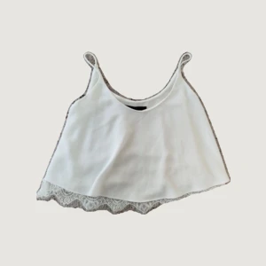 Vit linnetopp med spetskant - Söt vit linnetopp med tunna axelband och dekorativ spetskant längst ner. Toppen är ärmlös och har en luftig, croppad passform som passar perfekt till varma dagar. Enkel och stilren design som är lätt att matcha.