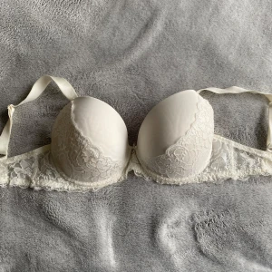 Vit spets-bh med rosett  - Supersöt vit bh med vadderade kupor och spetsdetaljer över hela bandet och kuporna. Framtill sitter en liten rosett med ett hjärtformat smycke. Aldrig använd. Storlek 75G och ingen push up. 