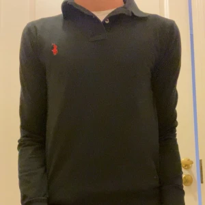 Svart pikétröja från Ralph Lauren - Klassisk svart långärmad pikétröja från Ralph Lauren med röd broderad logga på bröstet. Tröjan har krage, knappar vid halsen och är gjord i mjuk bomull. Perfekt för en clean och stilren look.