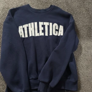 Mörkblå sweatshirt ATHLETICA Gina Tricot - Mörkblå sweatshirt från Gina Tricot med stor vit ATHLETICA-text framtill. Klassisk rund halsringning, ribbade muddar vid ärmslut och nederkant. Perfekt för en sportig och chill look. Mjuk insida och relaxed passform.