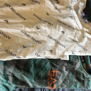 2 Emporio Armani t-shirts - Vit t-shirt från Emporio Armani med svart logotyp- och textprint över hela plagget. Klassisk rund halsringning och korta ärmar. Snygg och stilren design som sticker ut med det ikoniska Armani-mönstret. En militärgrön T-shirt från Emporio Armani, EA7 logga i orange. Strl S. Perfekt för dig som gillar exklusiva märken.