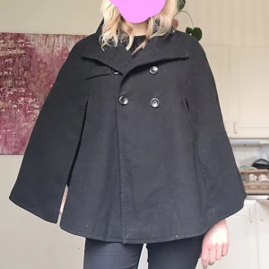 Svart cape med vida ärmar - Säljer en svart cape med snygg silhuett. Jackan har dubbelknäppning framtill och hög krage. Perfekt för dig som vill ha en stilren och trendig look! Så himla snygg men kommer tyvärr inte till så mycket användning 💕 öppen för prisförslag!