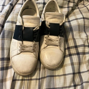 Valentino Garavani vit/svart - Snygga Valentino Garavani Open sneakers i vitt skinn med bred svart rem över vristen och coola nitar baktill. Klassisk rund tå, platt sula och vita. Några defekter på skorna sen ja la upp bilden så fråga så kan jag visa
