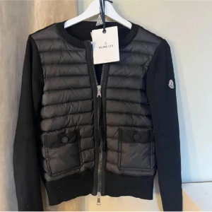 Moncler cardigan  - Svart pufferjacka från Moncler med quiltad front, två fickor med knappar och dragkedja. Ärmarna är släta och jackan har rund halsringning. Perfekt för dig som vill ha en stilren och sportig look. 