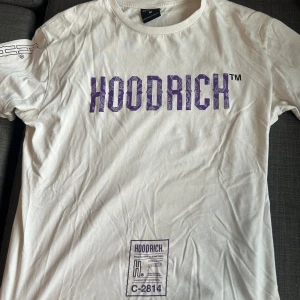 Vit t-shirt från Hoodrich med tryck - Vit t-shirt från Hoodrich med lila och svart tryck både fram och bak. Stort HOODRICH-tryck på bröstet och grafiska detaljer på ryggen. Klassisk rund hals och korta ärmar. Skön streetwear-vibe, perfekt till jeans eller shorts.