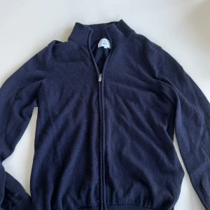 Mörkblå stickad cardigan från Bläck - Mörkblå stickad cardigan från Bläck med dragkedja framtill och hög krage. Långärmad och ribbade muddar vid ärmslut och nederkant. Perfekt för lager-på-lager och chill vardagsstil.