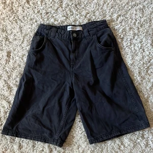 Svarta shorts från Bershka - Svarta, loose-fit shorts från Bershka med klassisk femficksdesign och bälteshällor. Längre modell som slutar strax ovanför knäna. Stängs med knapp och dragkedja framtill.
