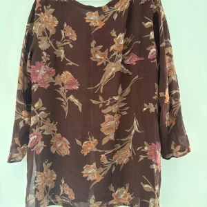 Blommig brun skjorta med knappar - Snygg brun skjorta med blommigt mönster i rosa, orange och beige toner. Skjortan har långa ärmar, knappar framtill och en loose passform. Perfekt för dig som gillar en avslappnad och bohemisk stil. Materialet känns lätt och luftigt. Står ej storlek men gissar på L-XL