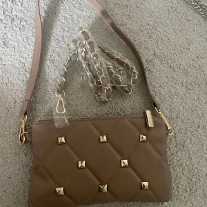 Valentino liknande väska med guldnitar - Snygg valentino liknande axelväska i quiltad design med guldiga nitar och dragkedja. Väskan har både axelrem i skinnimitation och en kedjerem med gulddetaljer. Insidan är fodrad med ljust mönstrat tyg och har flera fack för småsaker. Färg brun,beige, rosa färg 