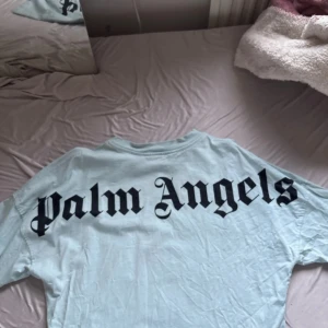Ljusblå Palm Angels t-shirt - Säljer en ljusblå t-shirt från Palm Angels med stor svart logga på ryggen och mindre logga vid halsen. Klassisk passform med korta ärmar och rund hals. Perfekt för dig som gillar streetwear och vill sticka ut med en clean look.