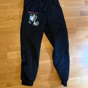 Svarta mjukisbyxor med tryck gymbyxor  - Svarta mjukisbyxor med elastisk midja och muddar vid bensluten. Snyggt tryck med motiv och röd text på ena benet ger en cool streetkänsla. Perfekta för chill eller häng med kompisar. Gymbyxor