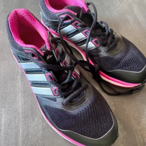 Svarta och rosa Adidas sneakers - Snygga Adidas sneakers i svart mesh och syntet med silvriga ränder och rosa detaljer. Skorna har svart snörning, rosa foder och vit/rosa sula. Perfekta för träning eller en sportig look. Klassisk Adidas-design med rund tå och platt sula.