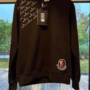 Svart Moncler sweatshirt XL - Svart sweatshirt från Moncler i storlek XL med tryckt logga över axeln och broderad patch nertill. Tröjan har rund hals, långa ärmar och ribbade muddar. Perfekt för dig som gillar stilrena och exklusiva plagg med tydlig branding.