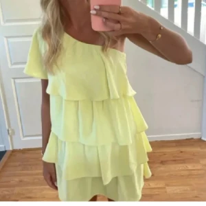 Gul volangklänning med oneshoulder - Superfin ljusgul kortklänning med flera volanger och oneshoulder-design. Klänningen har en luftig och flowy känsla, perfekt för varma dagar. Den är ärmlös på ena sidan och har en avslappnad passform som ger en cool och trendig look.