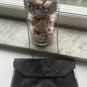 Svart kuvertväska med silverkedja - Stilren svart kuvertväska med glittrig yta och silverfärgade metalldetaljer. Väskan har en tunn kedja som axelrem och ett elegant kuvertlock. Perfekt när du vill addera lite extra bling till din outfit.